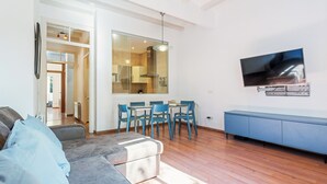 A modern living room with a grey sofa, a dining table with blue chairs, a TV mounted on the wall, and a wooden floor.