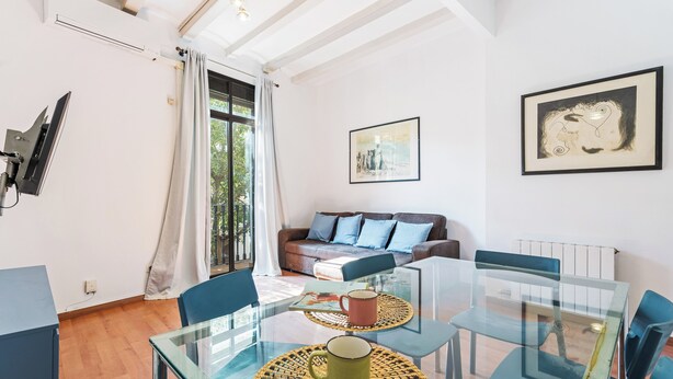 A modern living room with a glass dining table, blue chairs, a sofa with blue cushions, and a framed artwork on the wall.