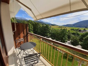 A balcony with a table and chairs, offering a view of a green valley and mountains.