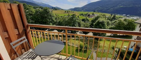 A balcony with a table and chairs, overlooking a mountainous landscape with greenery and buildings.
