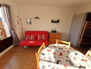 A room with a red sofa, a wooden dresser, a dining table with a deer pattern tablecloth, and a window with curtains.
