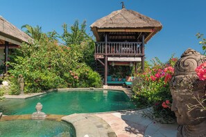 A pool area with a thatched-roof structure, lush greenery, and a stone sculpture. - Villa Kupu Kupu Tulip (Banjar)