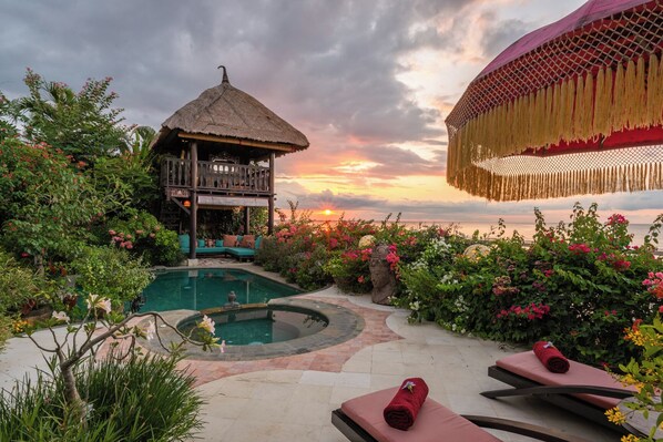 A pool area with a thatched-roof structure, lounge chairs, and a view of the sunset over the ocean. - Villa Kupu Kupu Tulip (Banjar)