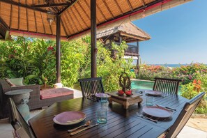 An outdoor dining area with wicker furniture, a table set for two, and a view of the ocean. - Villa Kupu Kupu Tulip (Banjar)