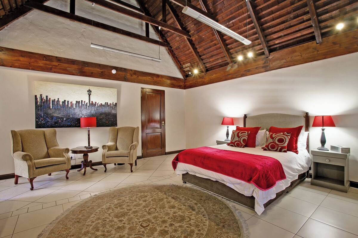Une chambre avec un grand lit, deux fauteuils, un tapis rond, un tableau représentant une vue panoramique d’une ville et un plafond en bois.