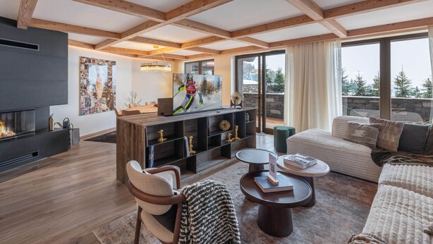 A modern living room with exposed wooden beams, a fireplace, a large TV, and a cozy seating area with a view of the outdoors.