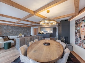A modern interior with a round wooden dining table surrounded by chairs, a stone wall, and a kitchen in the background.