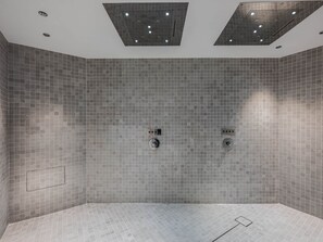Une salle de bain moderne avec des murs et un sol recouverts de carreaux gris, deux pommes de douche et des spots encastrés au plafond.