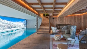 A modern indoor pool area with wooden beams, a view of snowy mountains, and a cozy seating area with patterned ottomans and wicker chairs.