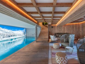 Un espace piscine intérieur moderne, agrémenté de poutres en bois, offrant une vue sur des montagnes enneigées, et comprenant un coin salon confortable avec des poufs à motifs et des fauteuils en osier.
