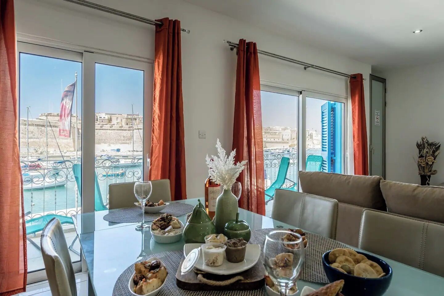 A dining area with a table set for two, a view of the harbor, and a balcony with chairs.