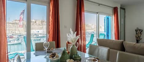 A dining area with a table set for two, a view of the harbor, and a balcony with chairs.
