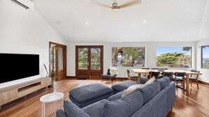 A modern living room with a large flat-screen TV, a sofa, a dining table with chairs, and a ceiling fan. - Chatby House filtered views of the ocean (Lorne)