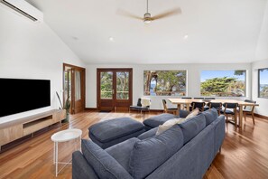 A modern living room with a large flat-screen TV, a sofa, a dining table with chairs, and a ceiling fan.