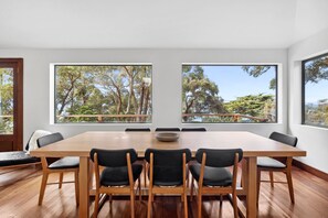 A modern dining room with a large wooden table, six chairs, and three large windows offering a view of trees and a balcony.