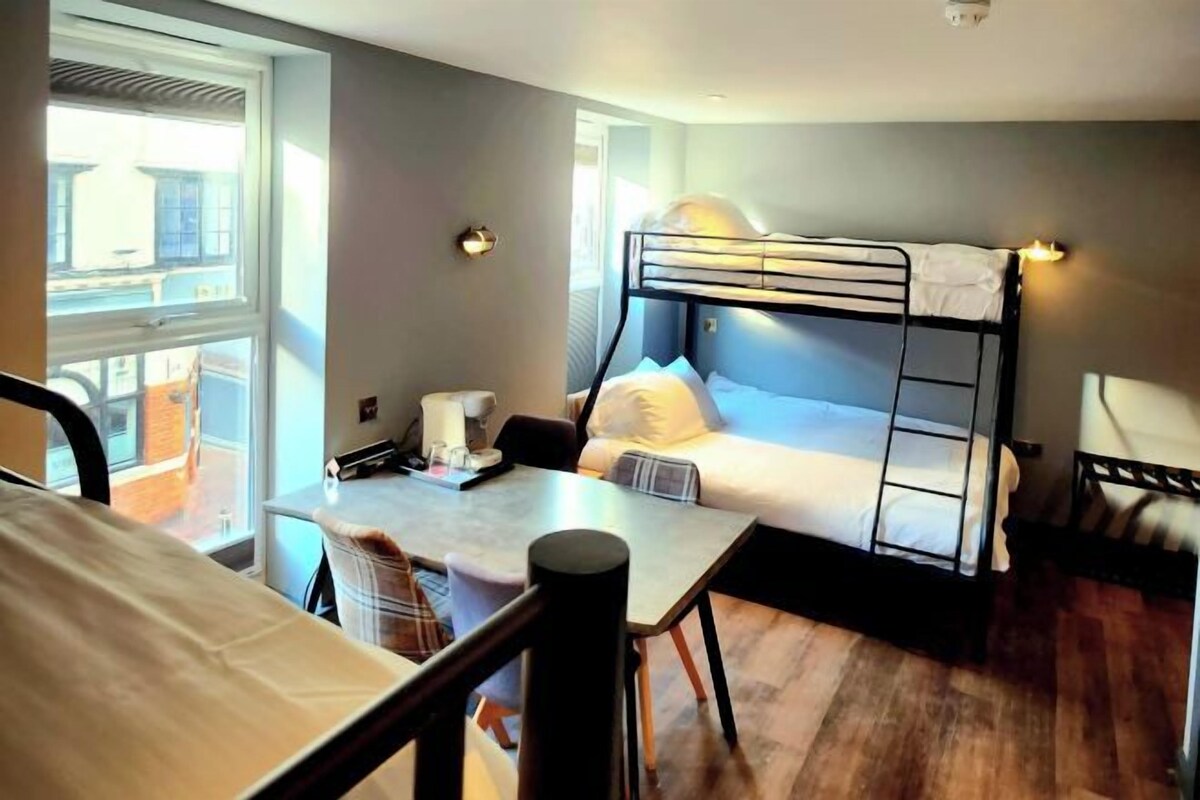 A room with a bunk bed, a desk, and a chair. There is a window providing natural light.