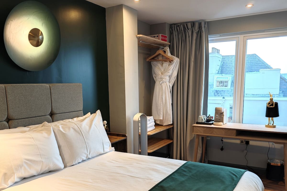 Habitación de hotel con una cama grande, una mesita de noche con lámpara, un perchero para bata y vistas a la ciudad a través de la ventana.