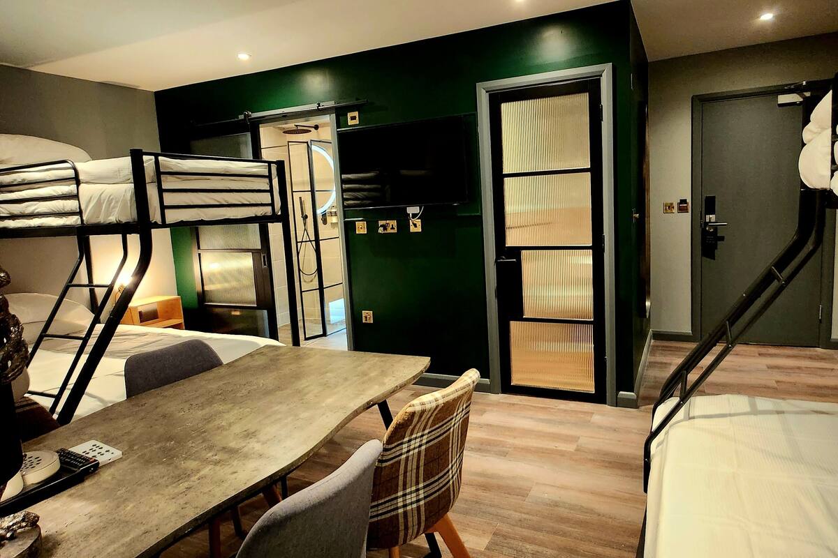 Habitación de hotel compacta con literas, una mesa de madera, sillas, un escritorio pequeño con una computadora y una pared verde decorativa.
