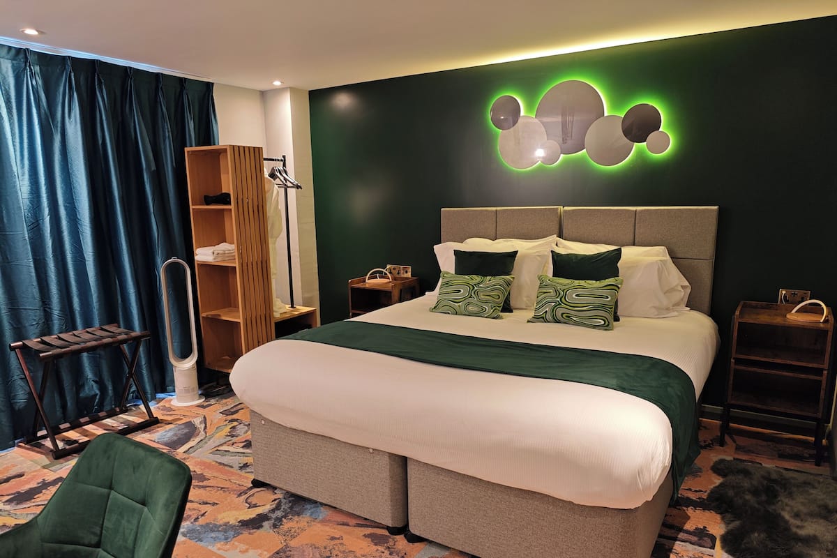 Una habitación de hotel moderna con una cama grande, un sillón verde, un armario de madera y una decoración de pared original.