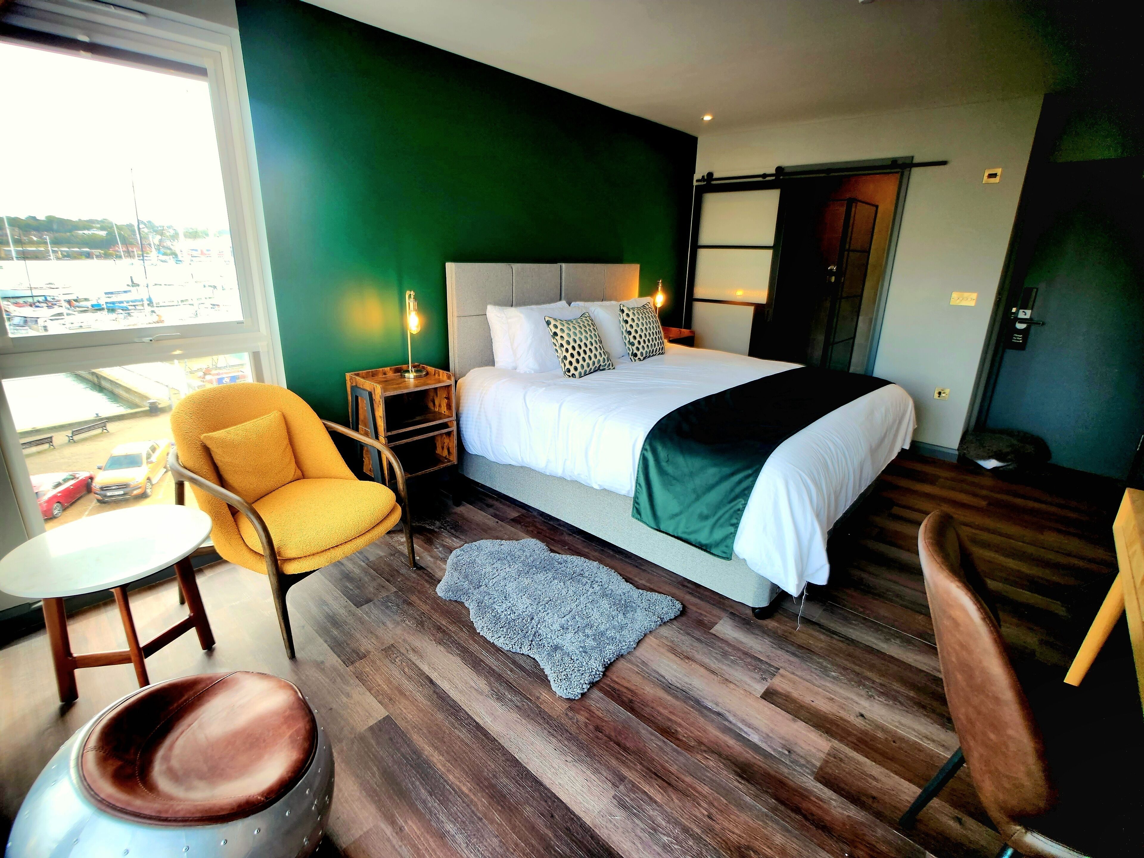 A modern hotel room with a large bed, a yellow armchair, a small round table, and a view of the outside.