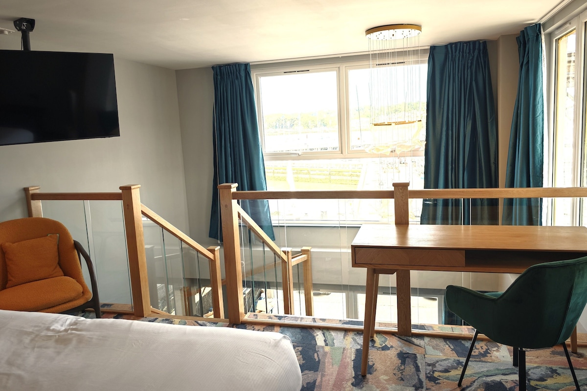 Habitación de hotel con cama, escritorio, silla, televisor, escalera y ventana con cortinas.
