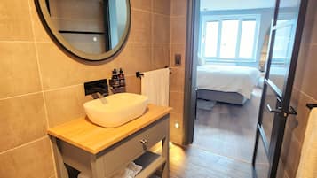 Un baño moderno con un espejo redondo, un lavabo blanco y un tocador de madera. Se ve una puerta que conduce a un dormitorio con cama y ventana.