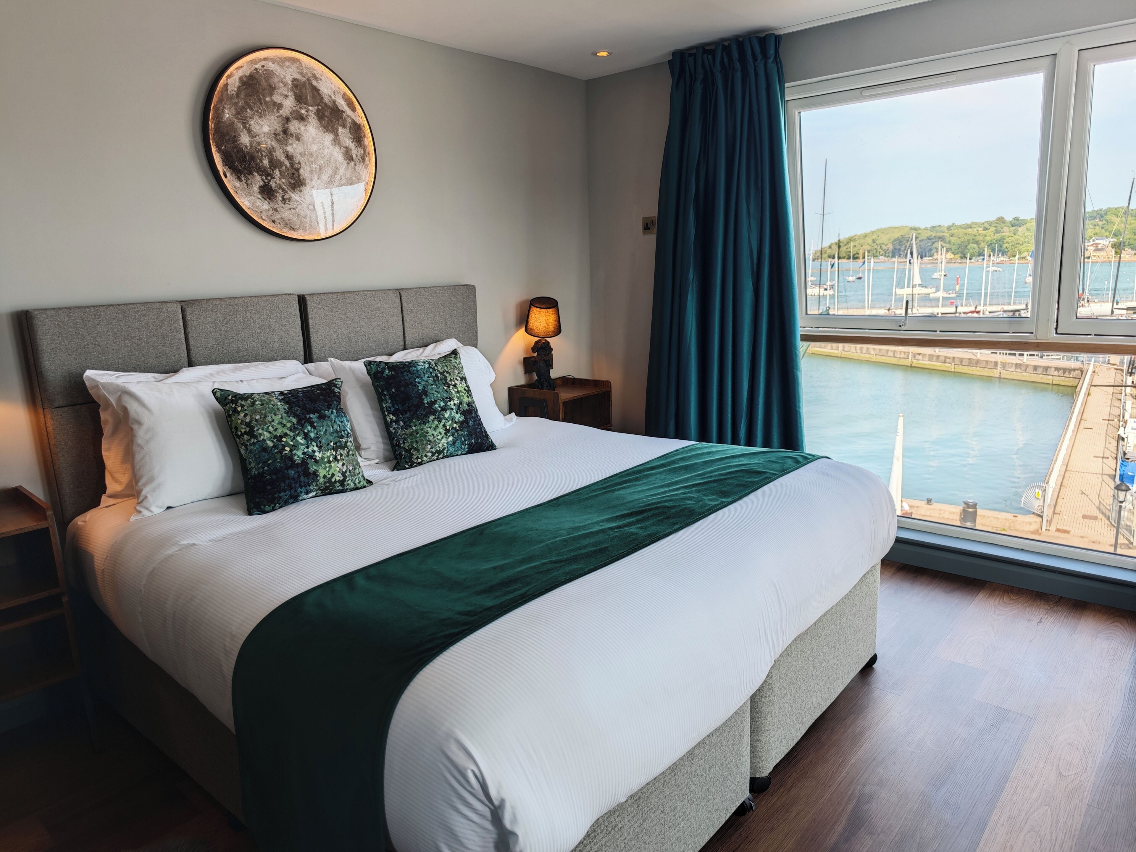 A bed with white and green bedding, a grey headboard, a nightstand with a lamp, and a view of a marina through a window.
