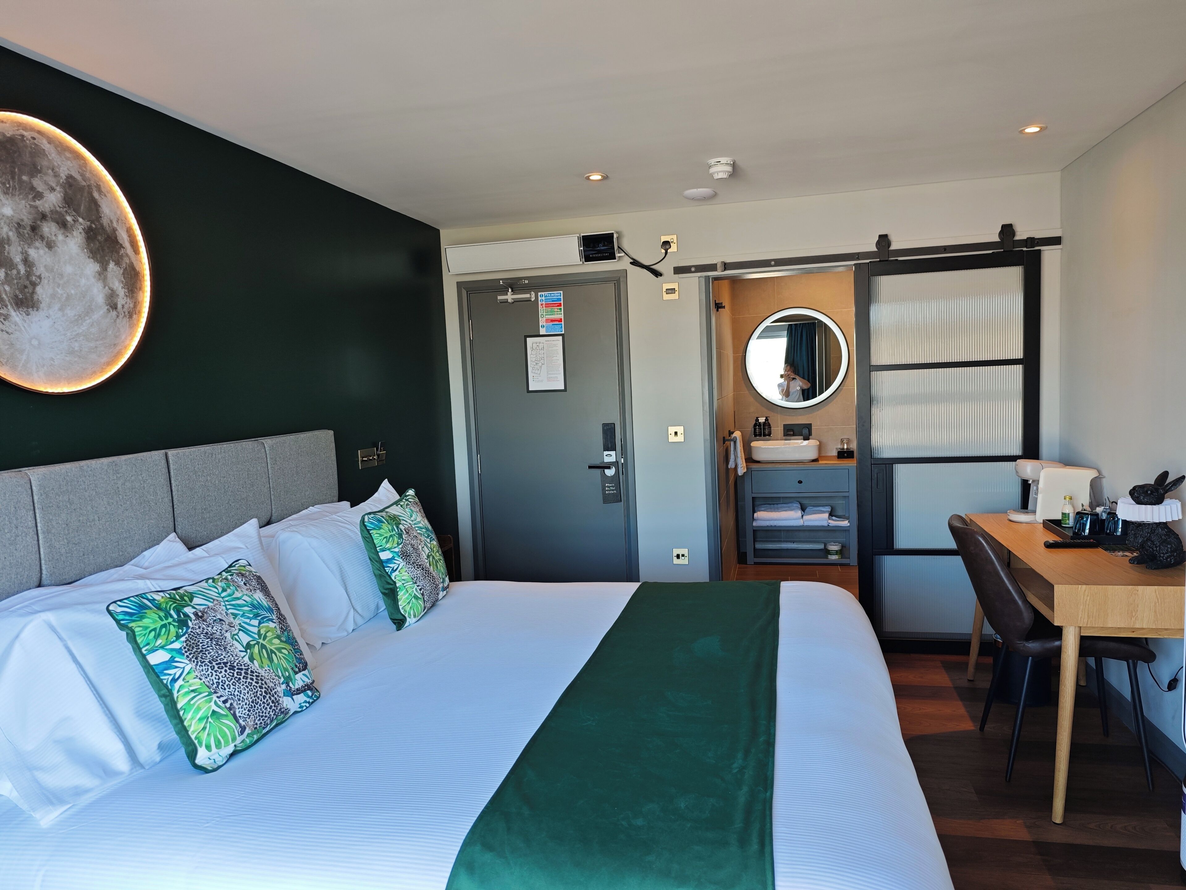 A hotel room with a bed, a desk, a chair, a mirror, and a bathroom with a sink and toilet.