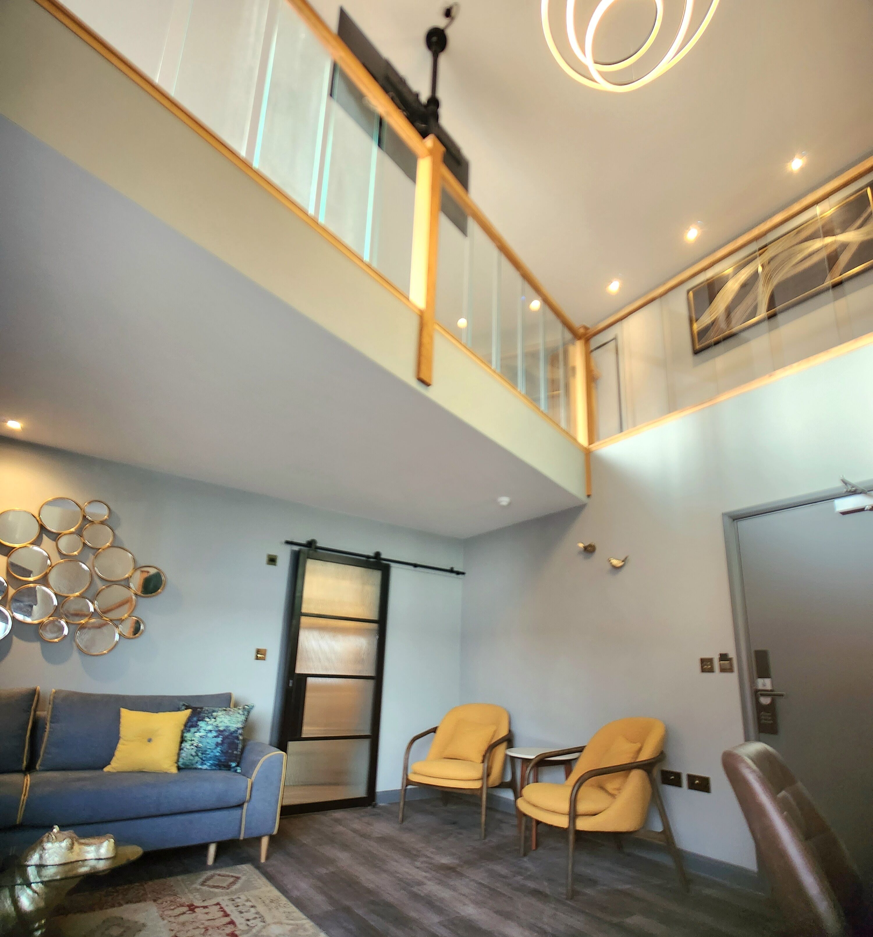A modern living room with a blue sofa, yellow chairs, a glass partition, and a staircase leading to an upper level.