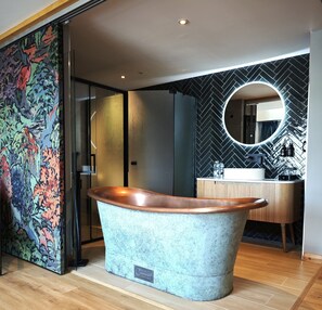 A modern bathroom with a freestanding copper bathtub, a round mirror, and a herringbone-patterned wall. - Pinnacle Suites (Cowes)
