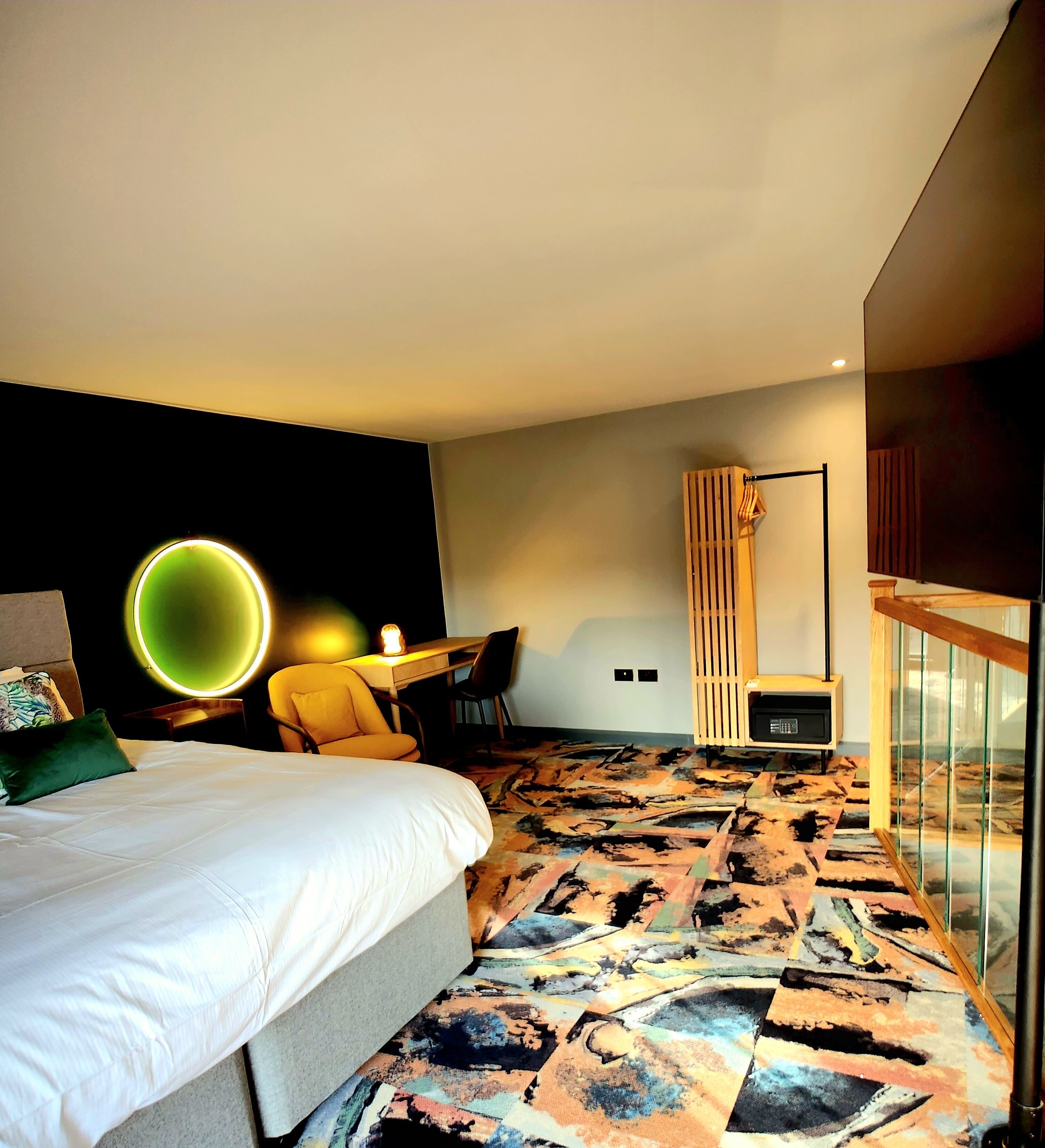 Quarto de hotel moderno com uma cama grande, uma mesa com cadeira, uma televisão pequena e um tapete com padrão único.