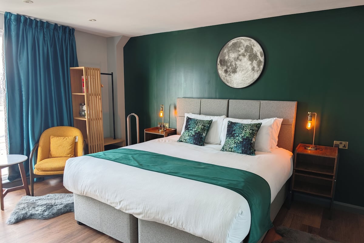 Una habitación de hotel moderna con una cama grande, dos mesitas de noche con lámparas, un atril, una silla, una estantería y un adorno de luna fijado a la pared.