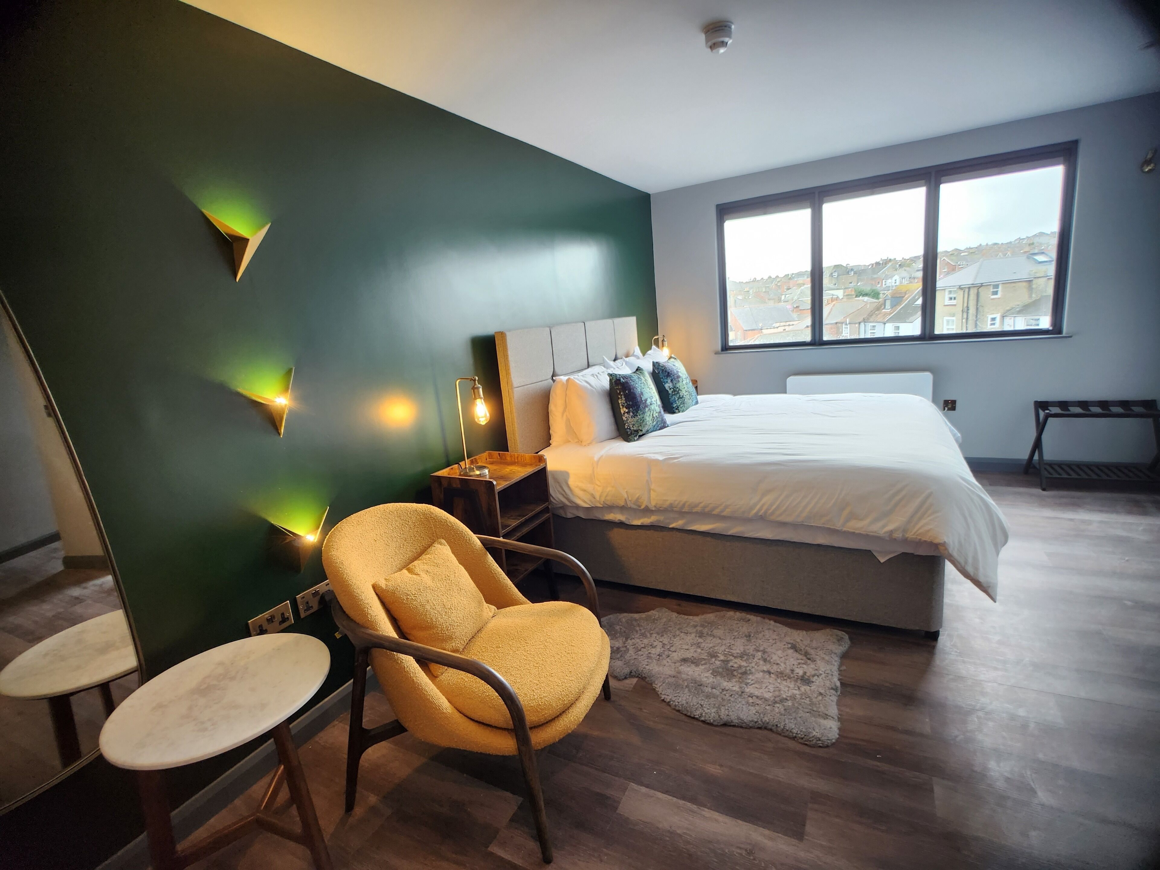Een moderne slaapkamer met een groot bed, een stoel, een klein tafeltje en uitzicht op de stad door een groot raam.