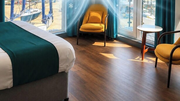A hotel room with a bed, a yellow armchair, a small round table, and a view of boats through a large window.