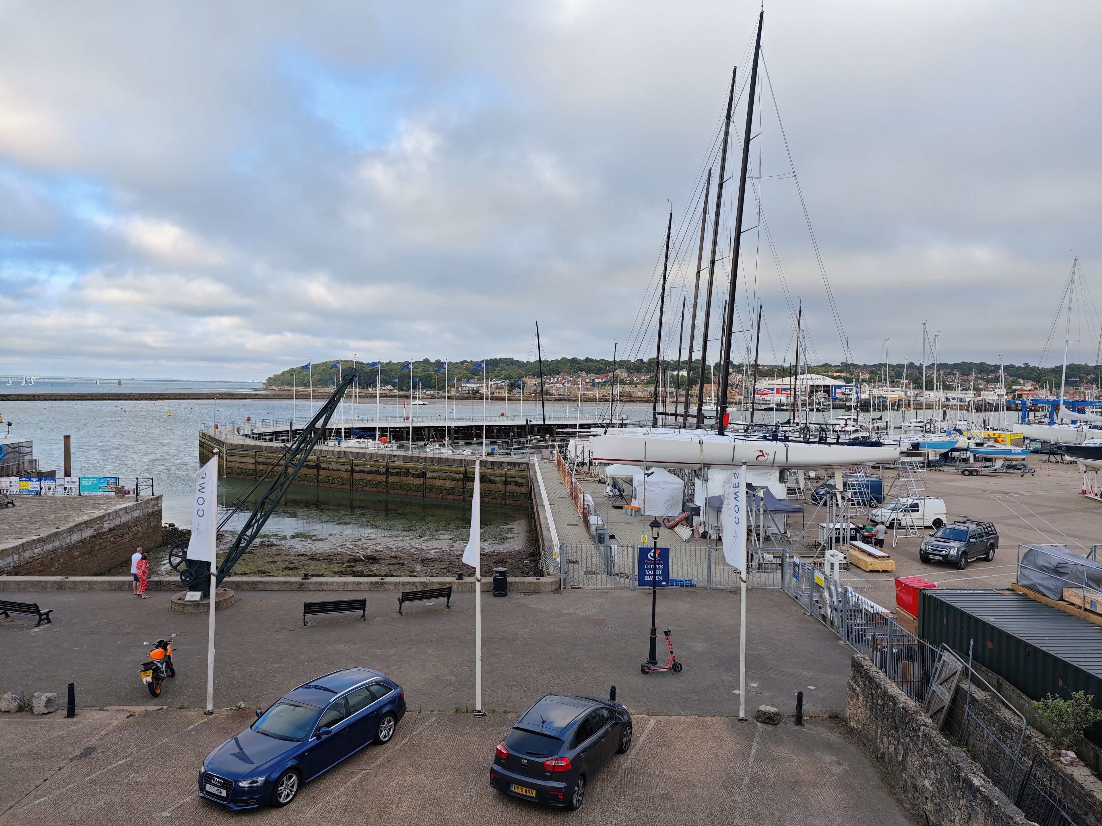 A marina with boats docked, a pedestrian walkway, and a parking area with cars and a scooter.