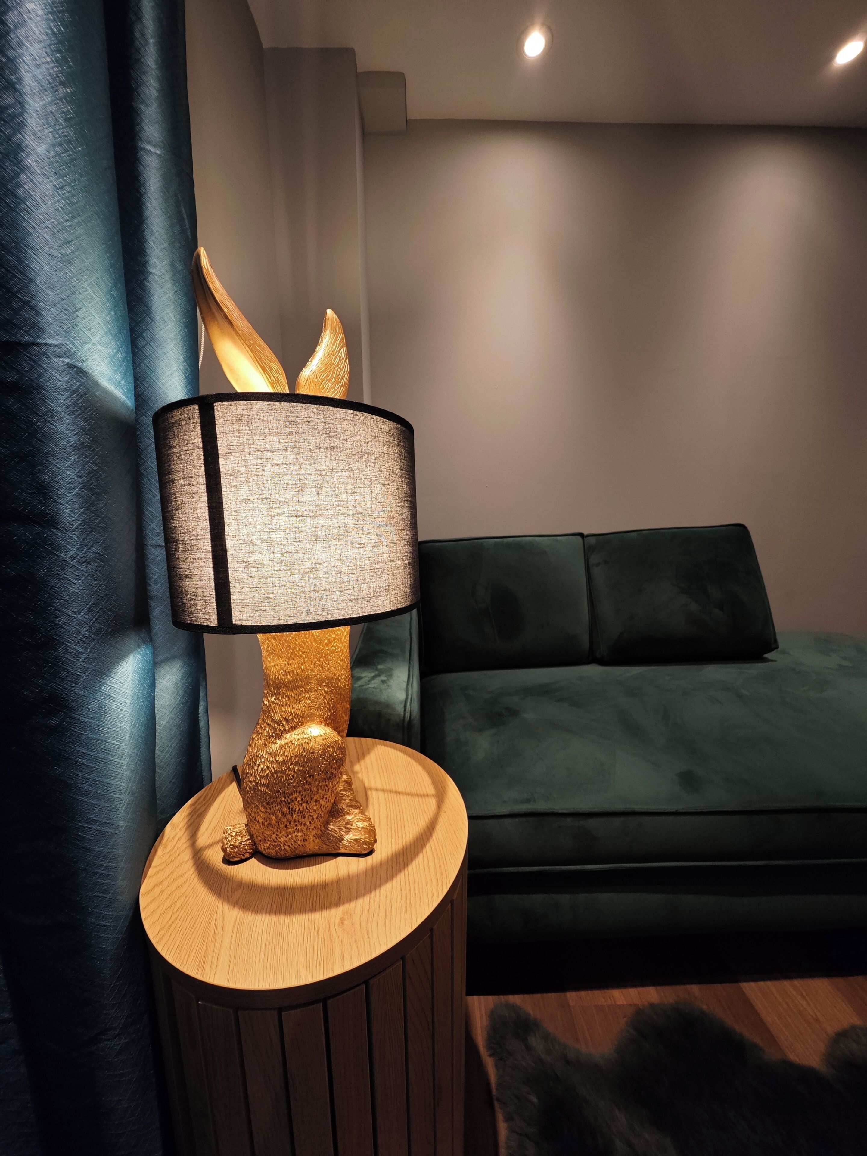 Ein modernes Wohnzimmer mit einem runden Holzhochentisch, einer Lampe mit strukturierter Schirm, einem grünen Sofa und einem dunklen Vorhang.