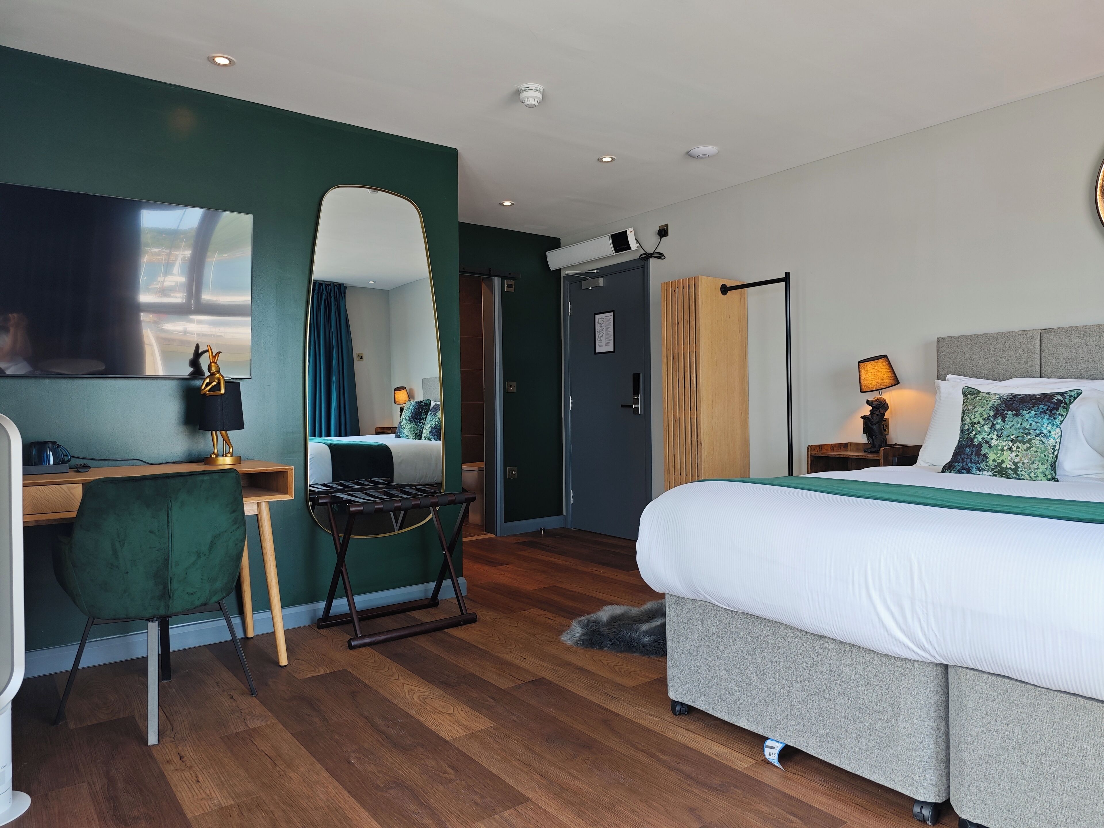 A modern hotel room with a large bed, a desk with a chair, a mirror, and a television.