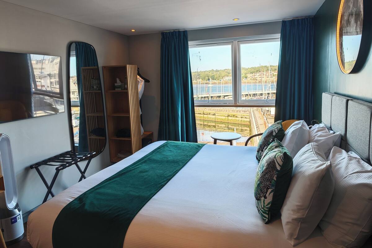 Una habitación de hotel con cama, una mesita, una silla, un armario y un ventanal con vista a un muelle y al agua.