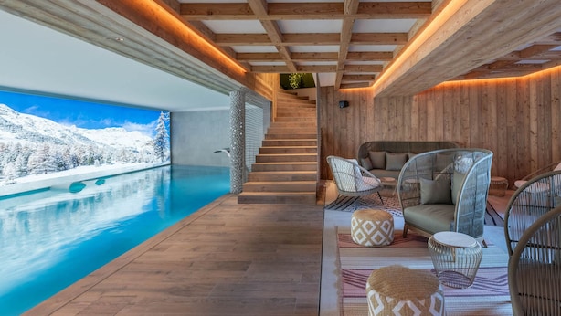 Ein moderner Innenpoolbereich mit Holzbalken, Blick auf verschneite Berge und eine gemütliche Sitzecke mit gemusterten Kissen.