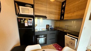A compact kitchen with a microwave, refrigerator, oven, and dishwasher. There is a dining table with chairs, a wooden cabinet, and a shelf with decorative items. - Lovely 2-room apartment, south balcony, mountain view, WIFI, 4 pax (Villard-de-Lans)