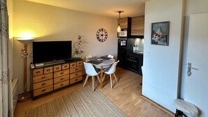 A compact living space with a TV on a wooden cabinet, a dining table with chairs, a clock on the wall, and a kitchen area with appliances. - Lovely 2-room apartment, south balcony, mountain view, WIFI, 4 pax (Villard-de-Lans)