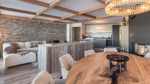 Amplia sala de estar con pared de piedra, vigas de madera, una gran mesa de comedor de madera y una cocina moderna al fondo.