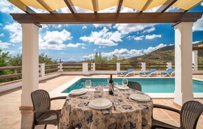 Relax by the pool surrounded by lush hills and greenery, with a pergola offering shade and a dining area nearby.