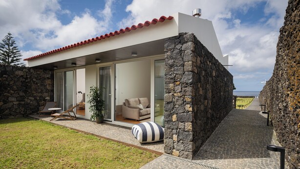 A modern house with a stone exterior, a red-tiled roof, and a glass door leading to a living area with a sofa and a potted plant. - ENTRE MUROS (Ribeira Grande)