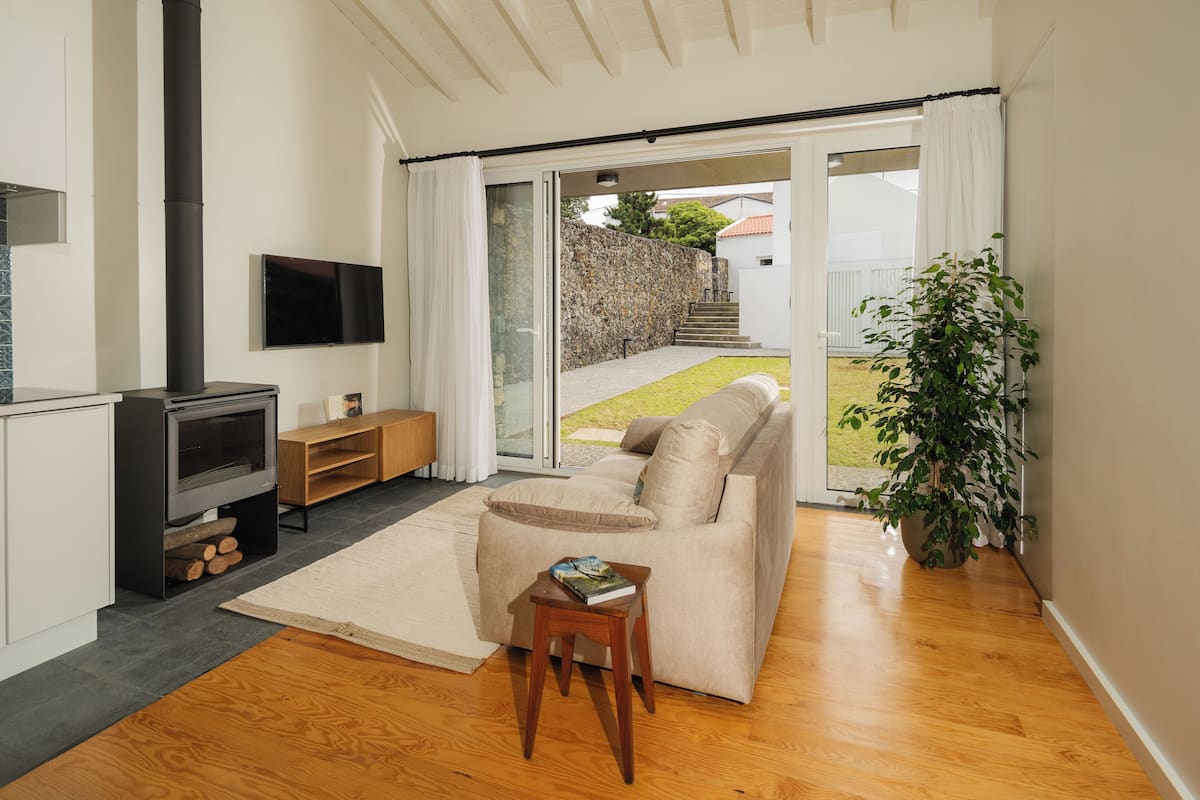 A modern living room with a sofa, a small wooden table with a book, a fireplace, and a large window overlooking a garden.