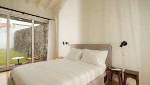 A bedroom with a large bed, a small table, a nightstand, and a view of a stone wall and garden through a sliding door. - ENTRE MUROS (Ribeira Grande)