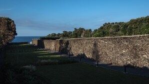 A stone wall with a grassy area in front, a clear sky, and trees in the background. - ENTRE MUROS (Ribeira Grande)