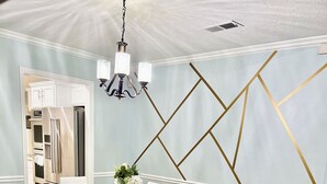 Un comedor con mesa y sillas blancas, un tapete estampado y una pared con detalles geométricos dorados.