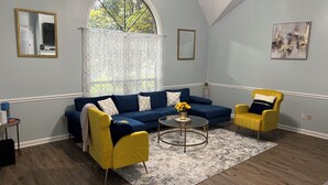 Una sala de estar con un sofá azul, sillones amarillos, una mesa de centro y un ventanal con vistas a árboles.