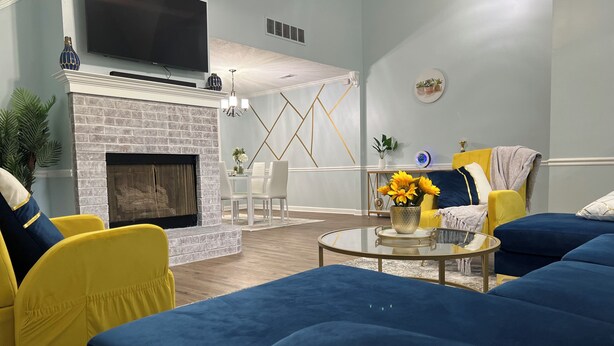 Una sala de estar moderna con chimenea, sillas amarillas, sofás azules y un televisor de pantalla plana montado sobre la chimenea.